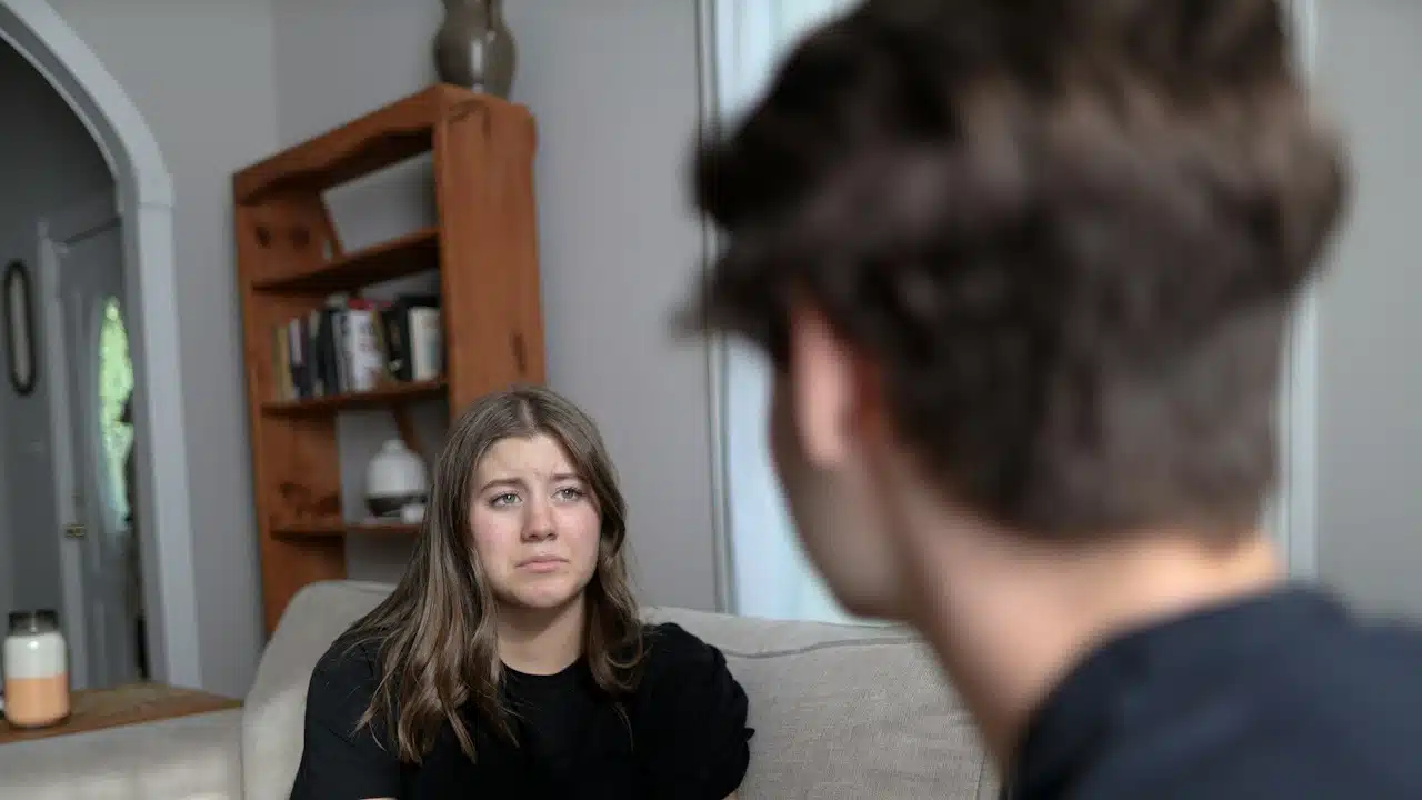 A woman looking at her husband while he is complaining about her behavior while they are sitting on a couch together.
