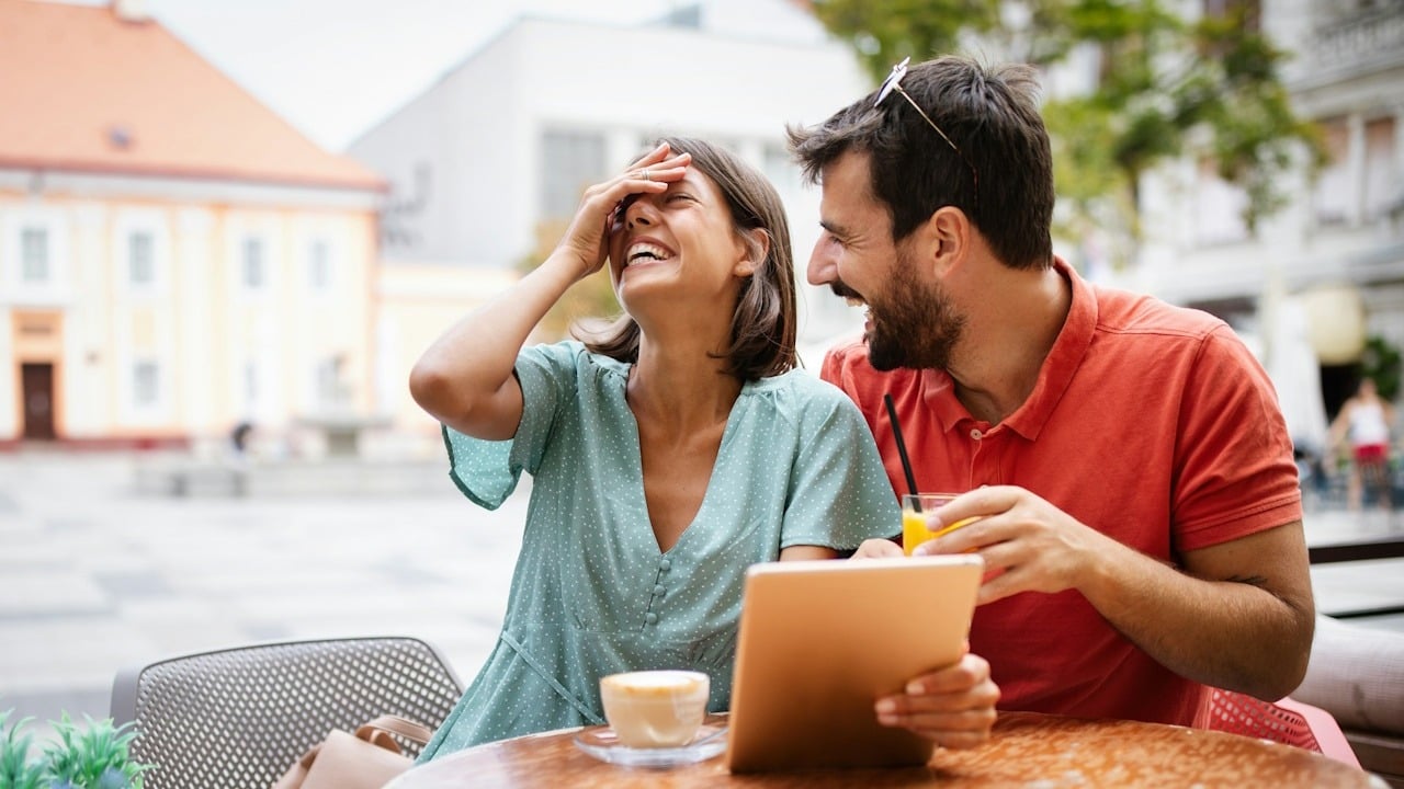 A couple laughing while sitting at a table and looking at a tablet.