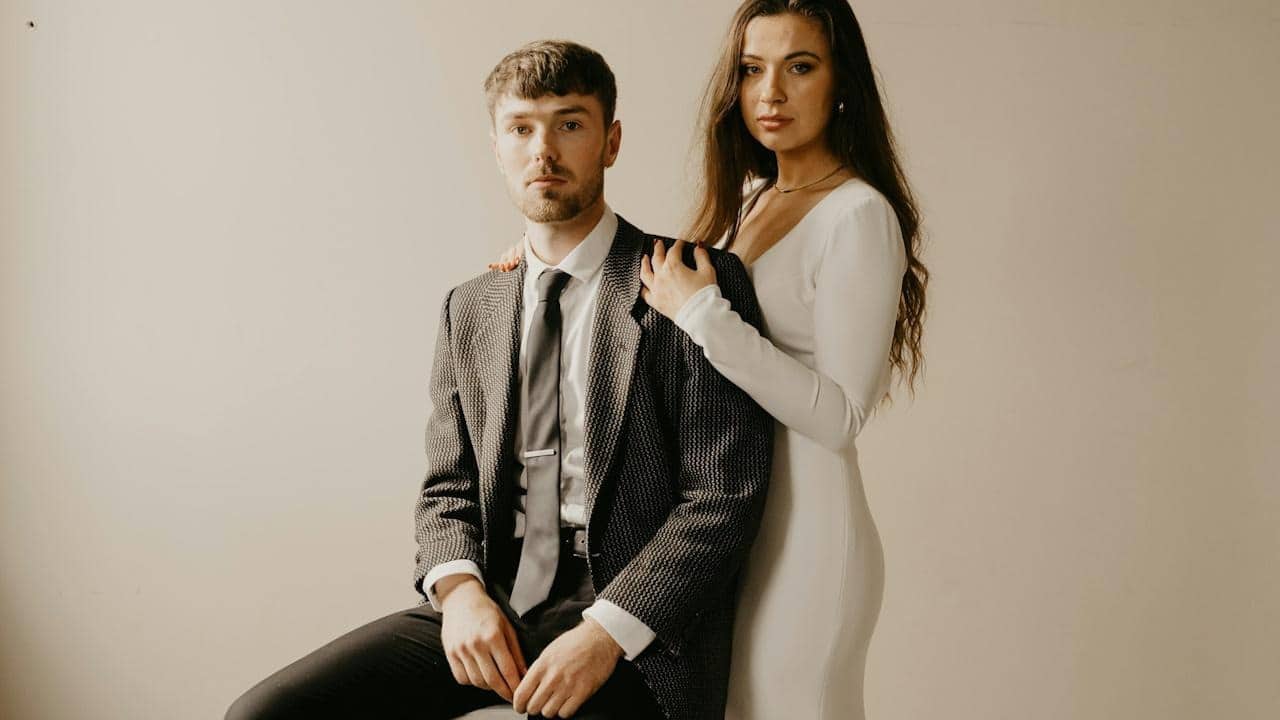 A woman resting her hands on a sitting man’s shoulders while she stands behind him.