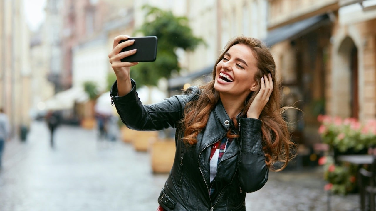 A woman smiling and taking a selfie while standing in a street and adjusting her hair.