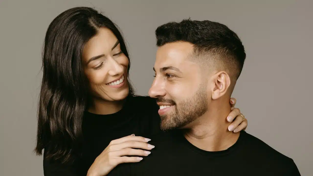 A woman is smiling at a lightly bearded man as he looks at her while she holds his neck lightly.