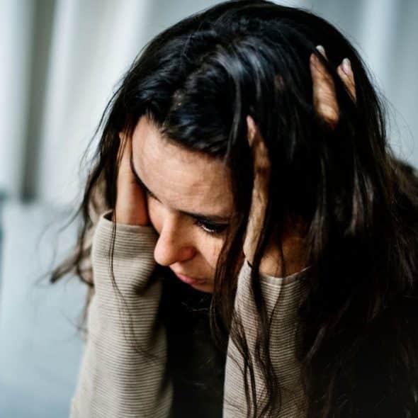 A woman with long dark hair holds her head in her hands while looking down.