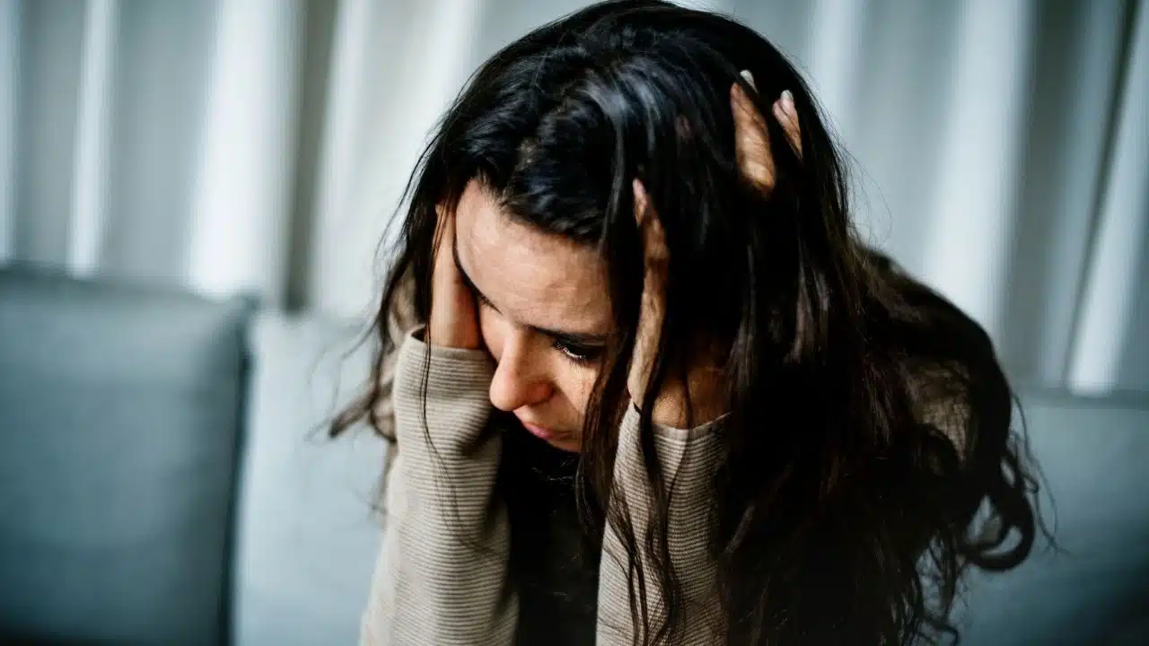 A woman with long dark hair holds her head in her hands while looking down.
