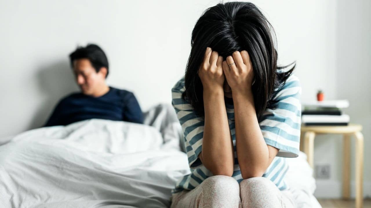 A woman sits on a bed with her face in her hands while a man sits behind.