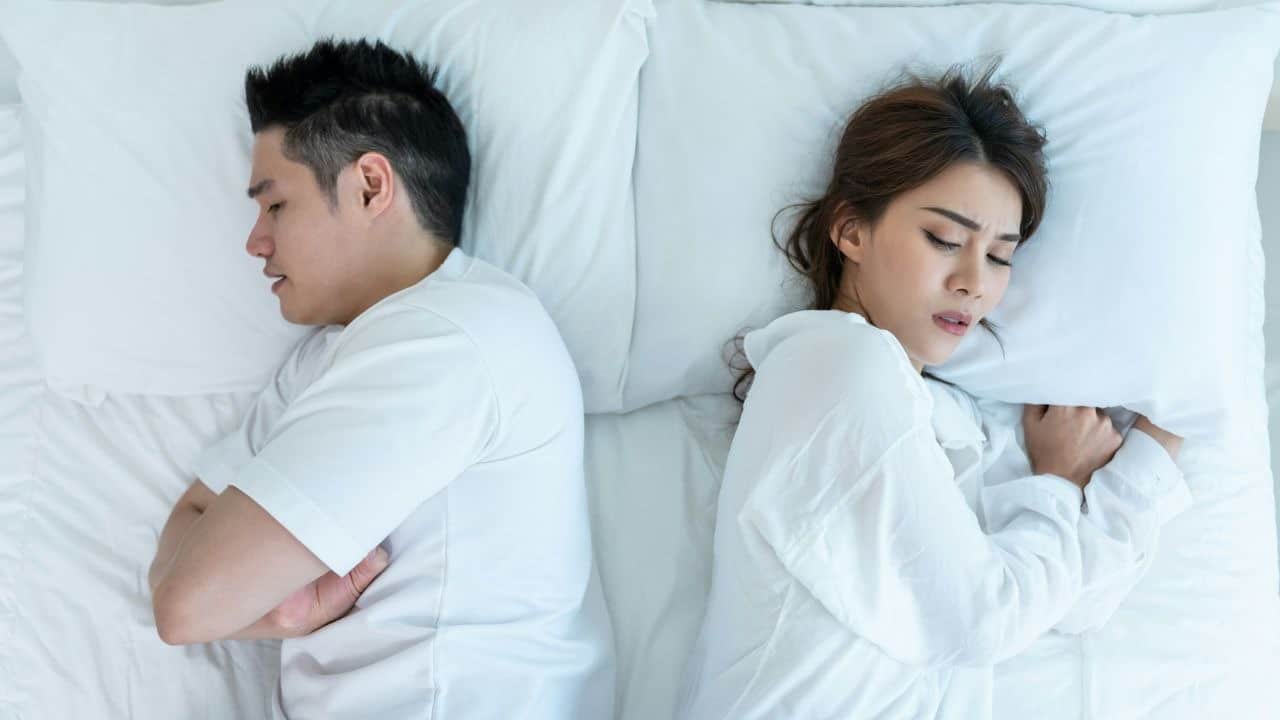 A man and woman lie in bed facing away from each other with eyes closed.