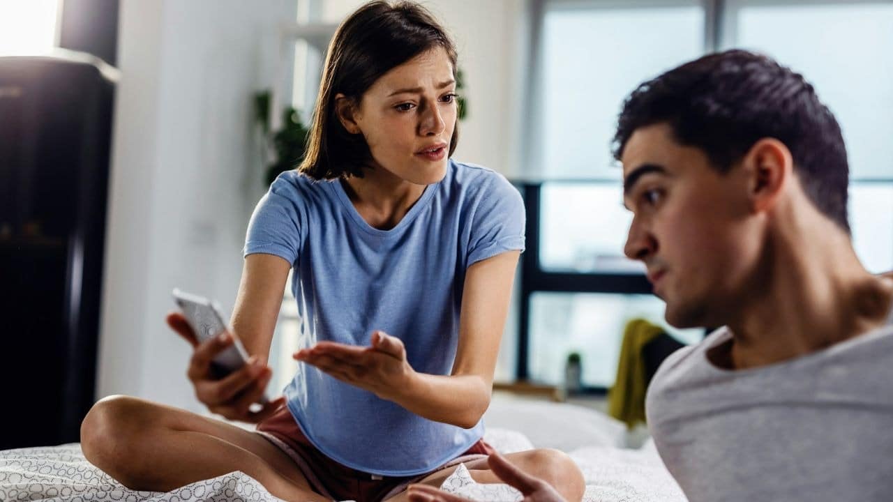 A woman holds a smartphone and gestures toward a man who is looking at her.
