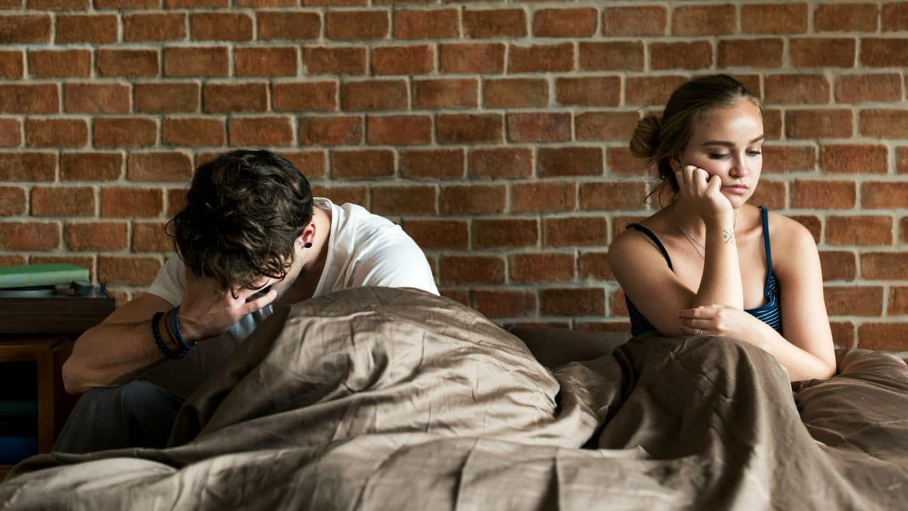 A man sits on a bed with his head in his hands near a somber woman.