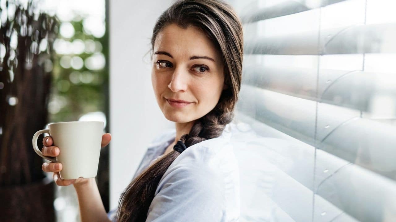 A woman with a long braid holds a white mug and looks out of a window.