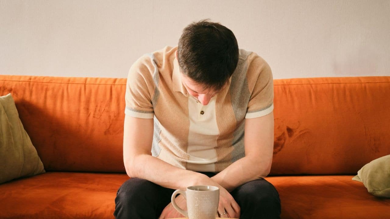 A man in a striped shirt sits on an orange sofa with his head bowed down.