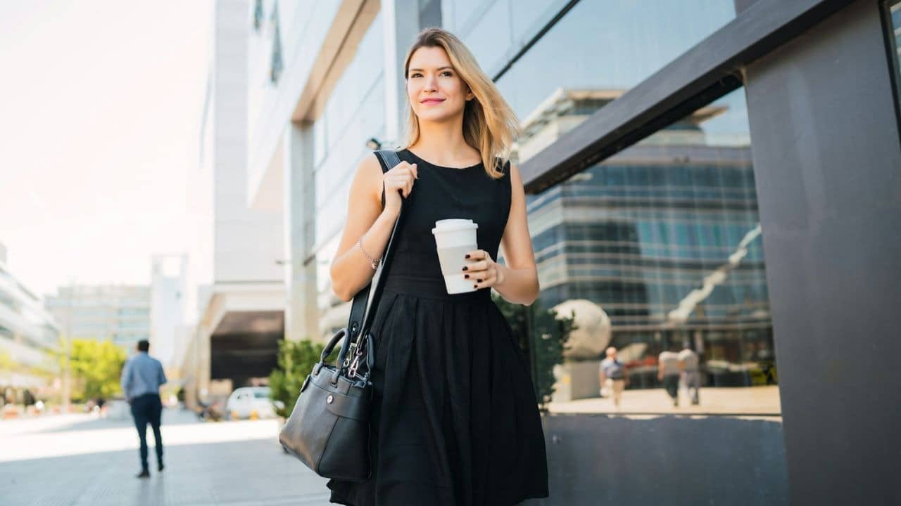 A woman in a black dress walks outside while carrying a cup and a bag.