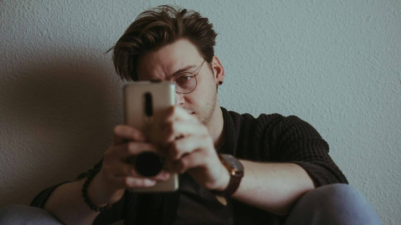 A man with glasses sits against a wall, looking intently at a smartphone held in both hands.