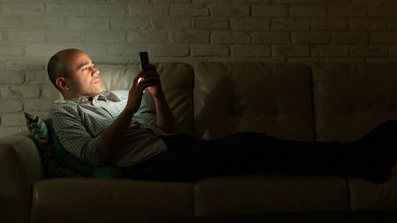 A man lies on a couch in a dark room, illuminated by the screen of his smartphone.