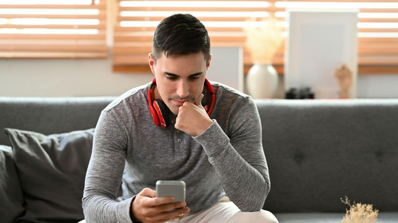 A man with red headphones around his neck sits on a couch looking at a smartphone.
