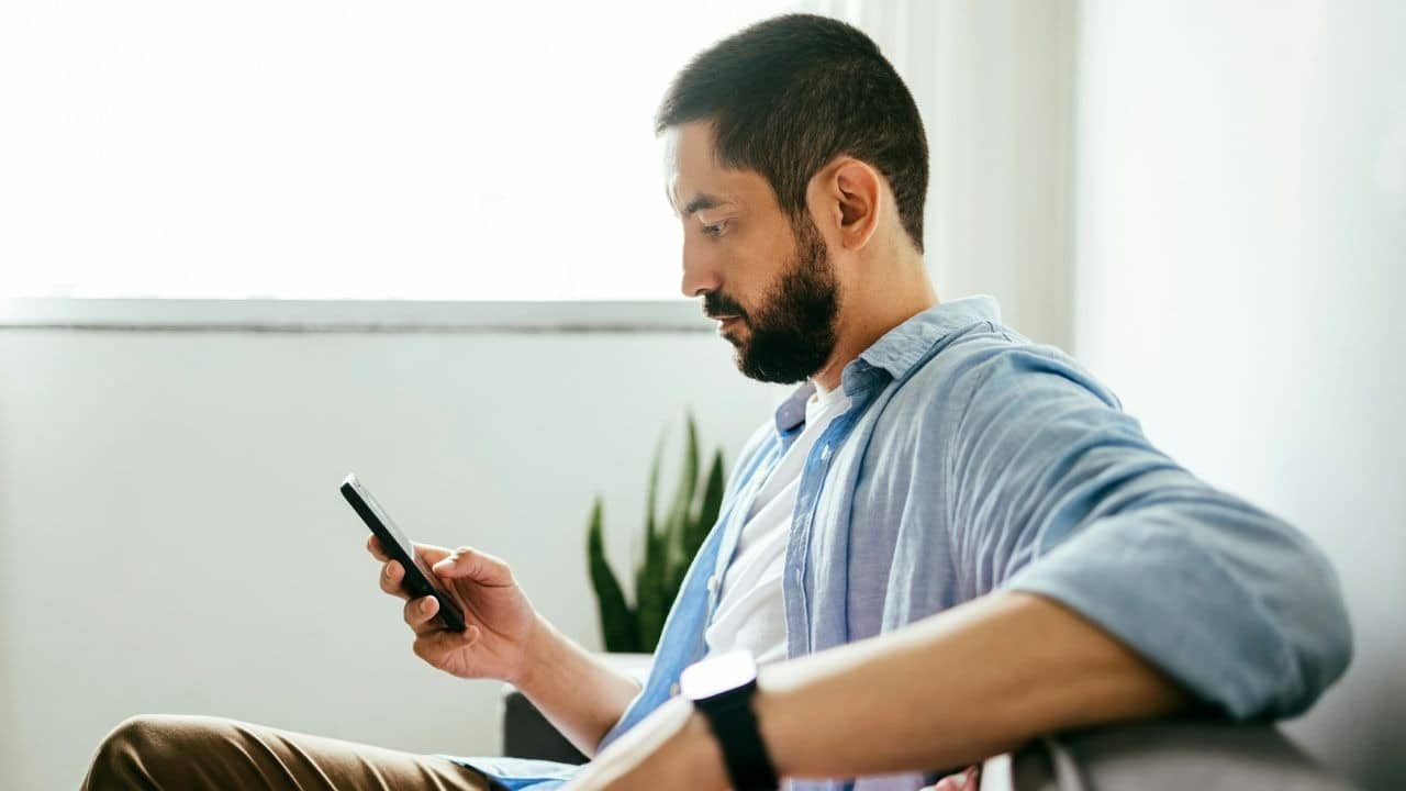 A bearded man in a blue button-down shirt sits indoors while looking at a handheld smartphone.