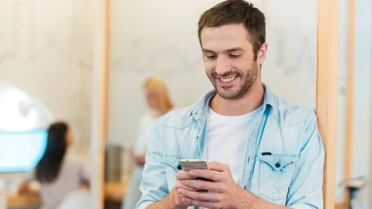 A smiling man in a light blue shirt leans against a doorway while using a smartphone.
