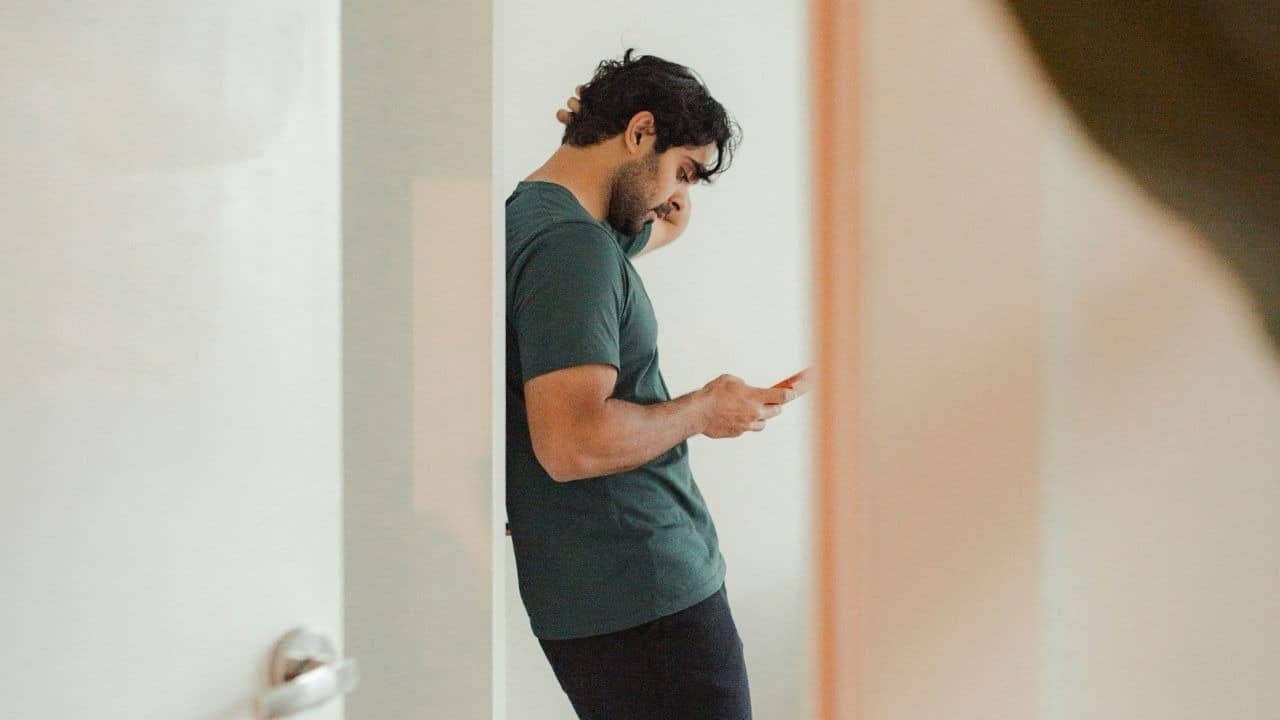 A man in a green t-shirt leans against a white wall while looking at his smartphone.