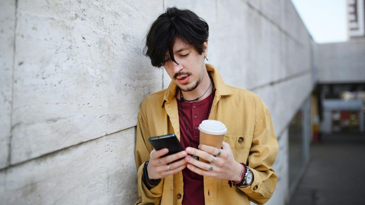A man in a yellow shirt leans against a wall holding a coffee cup and smartphone.