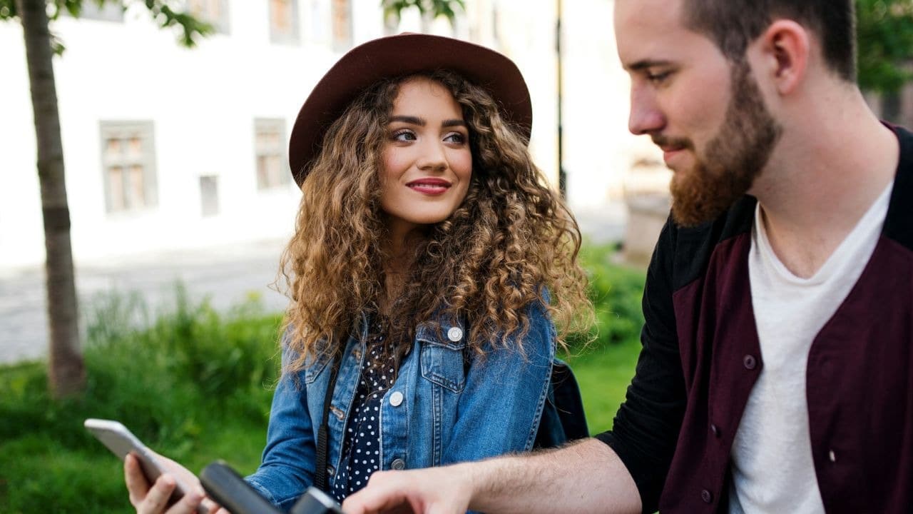 A woman with curly hair looks at a man while he holds a smartphone outdoors.