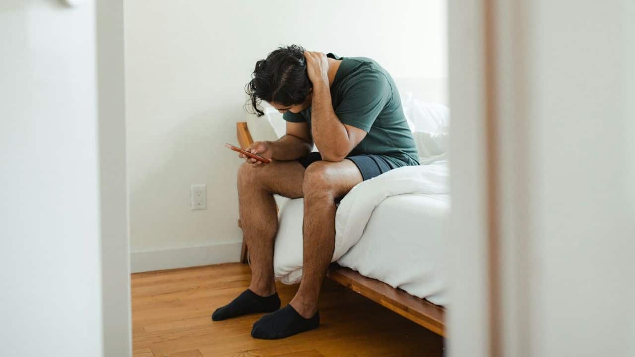A man in a green shirt sits on a bed, holding his head and looking down.