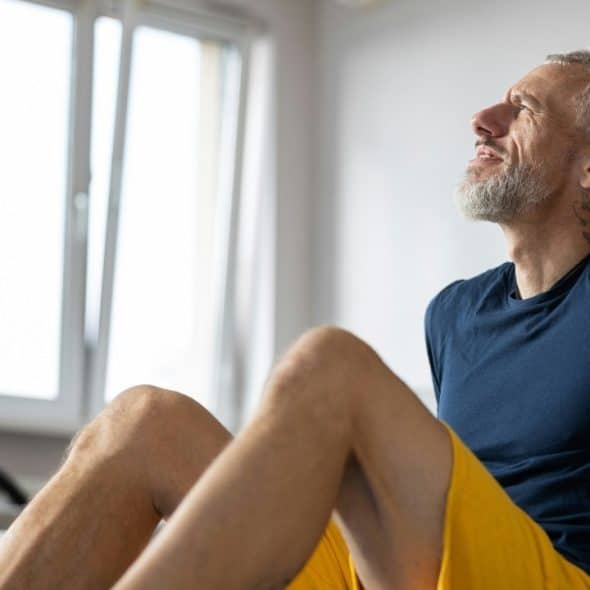 A bearded man in athletic wear sits indoors, looking upward toward a bright window.
