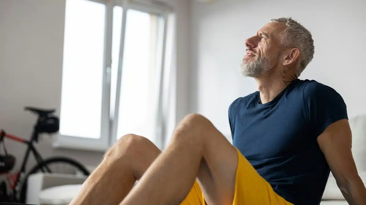A bearded man in athletic wear sits indoors, looking upward toward a bright window.