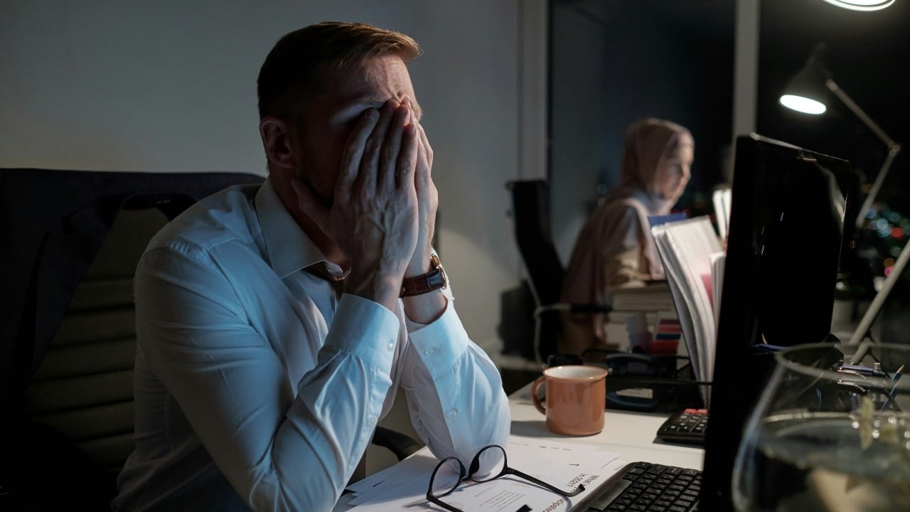 A man at a desk covers his face with both hands in a dark office.