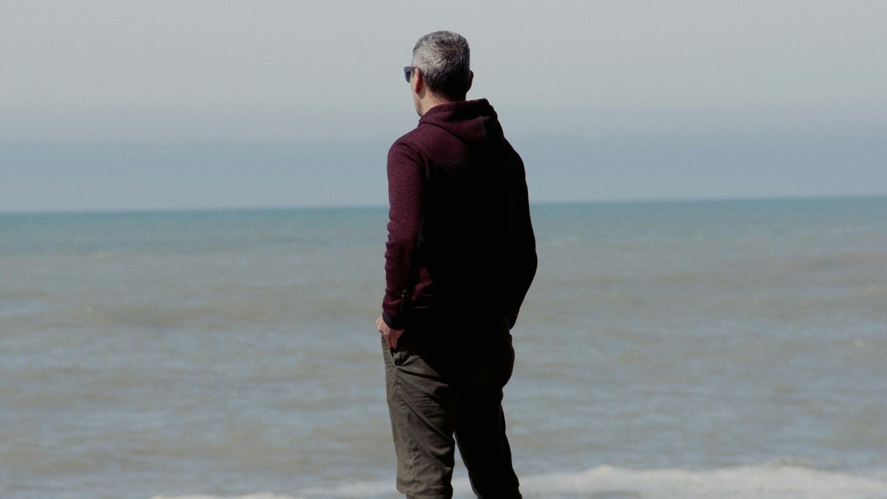 A man in a dark hoodie stands with his back to the camera, facing the ocean.