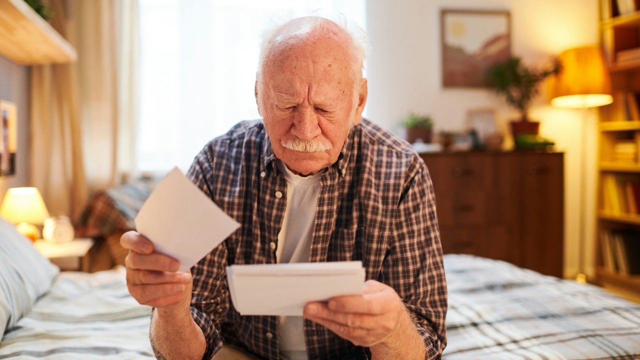 Comparing Yourself to Your Younger Self An elderly man with a mustache sits on a bed looking at several physical photographs.