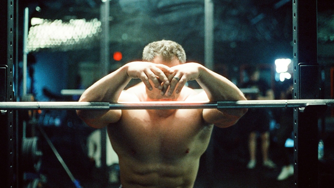 A muscular man rests his arms on a metal barbell inside a dimly lit gym.