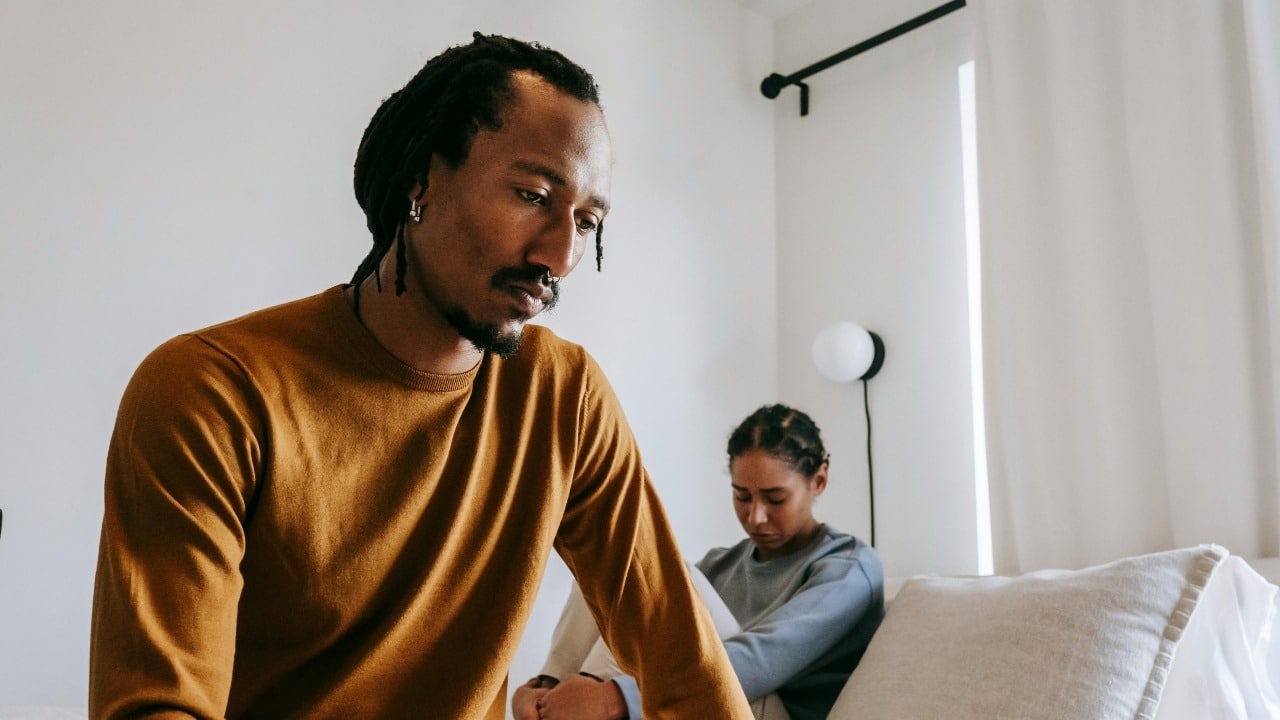 A man with dreadlocks looks down while a woman sits blurred in the background.