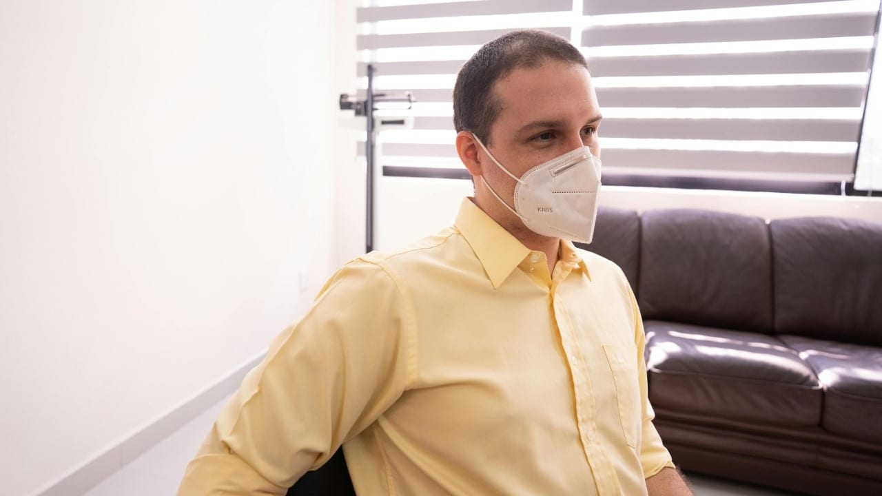 A man wearing a yellow shirt and a white face mask sits in an office.