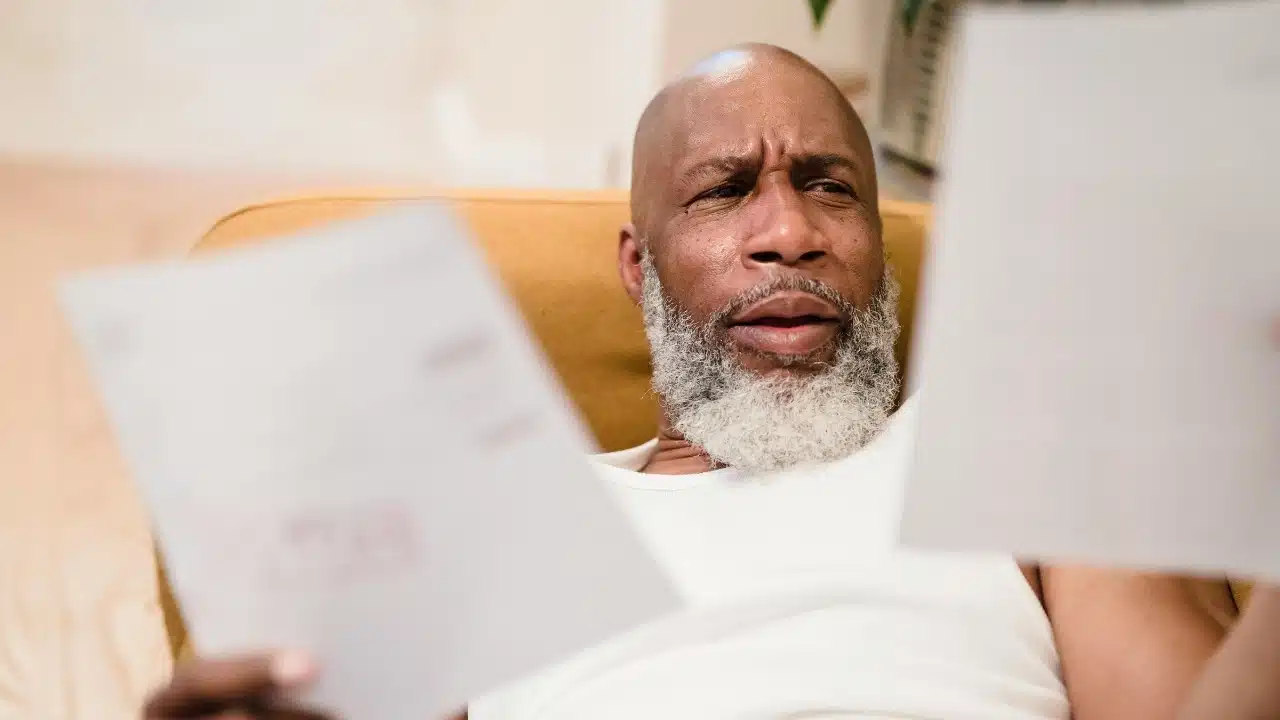 A man with a white beard holds papers and looks at them with a furrowed brow.