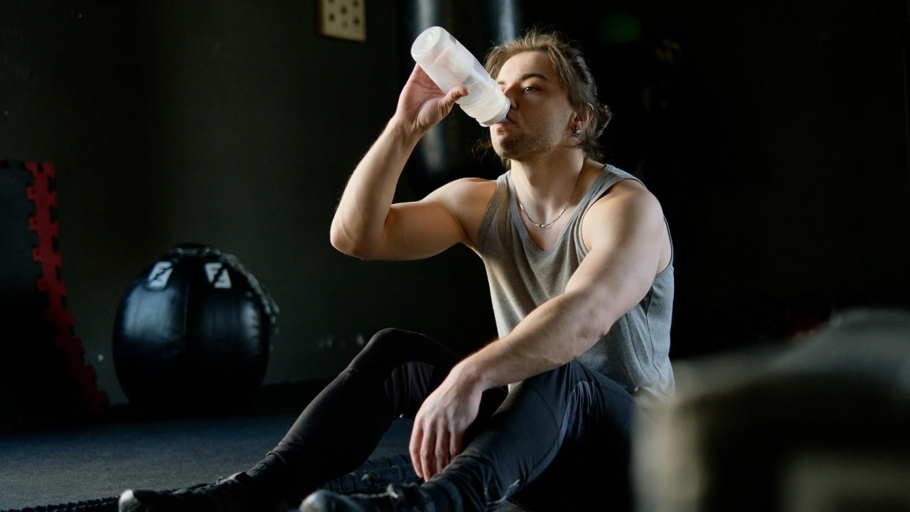 A man in a grey tank top sits on a gym floor drinking from a bottle.