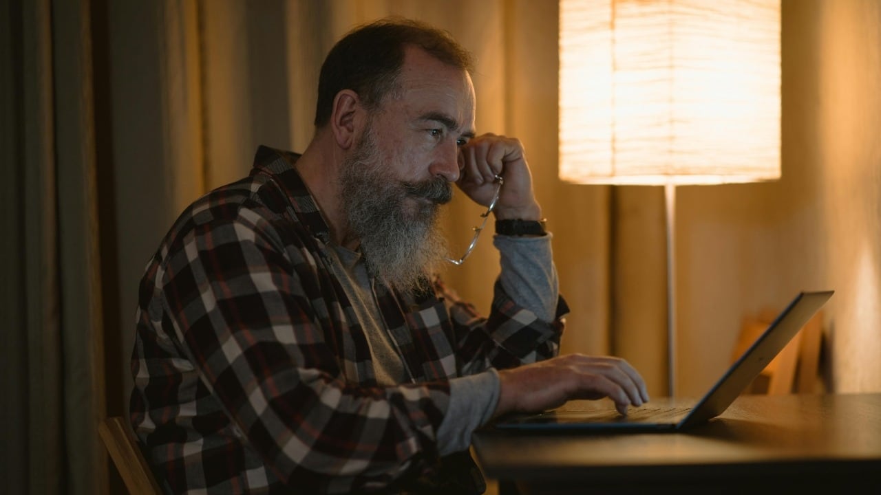 A man with a long grey beard types on a laptop near a glowing lamp.