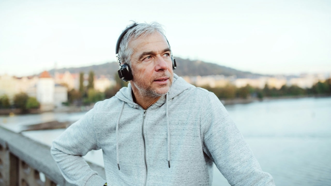 A man wearing a grey hoodie and headphones looks out over a body of water.