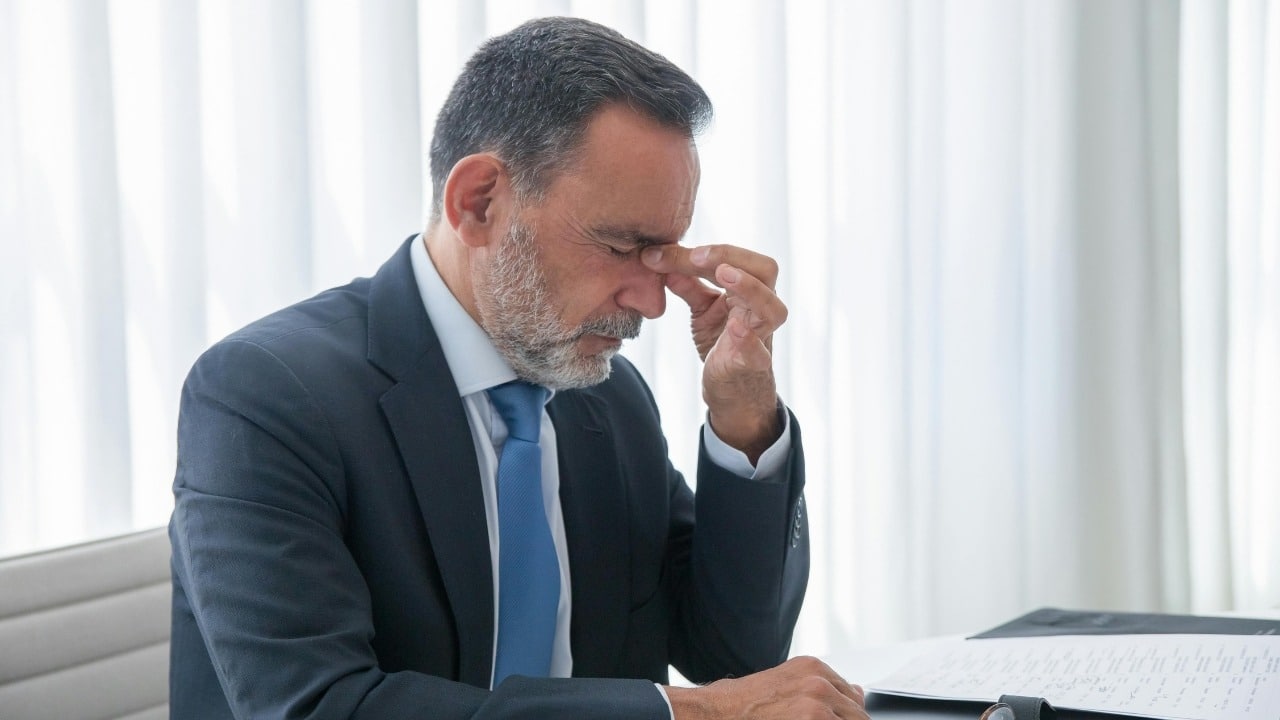 A man in a suit and blue tie pinches the bridge of his closed eyes.