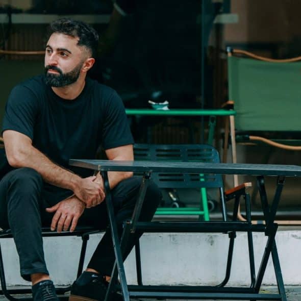 Bearded man in a black t-shirt sits at an outdoor table looking to the side.