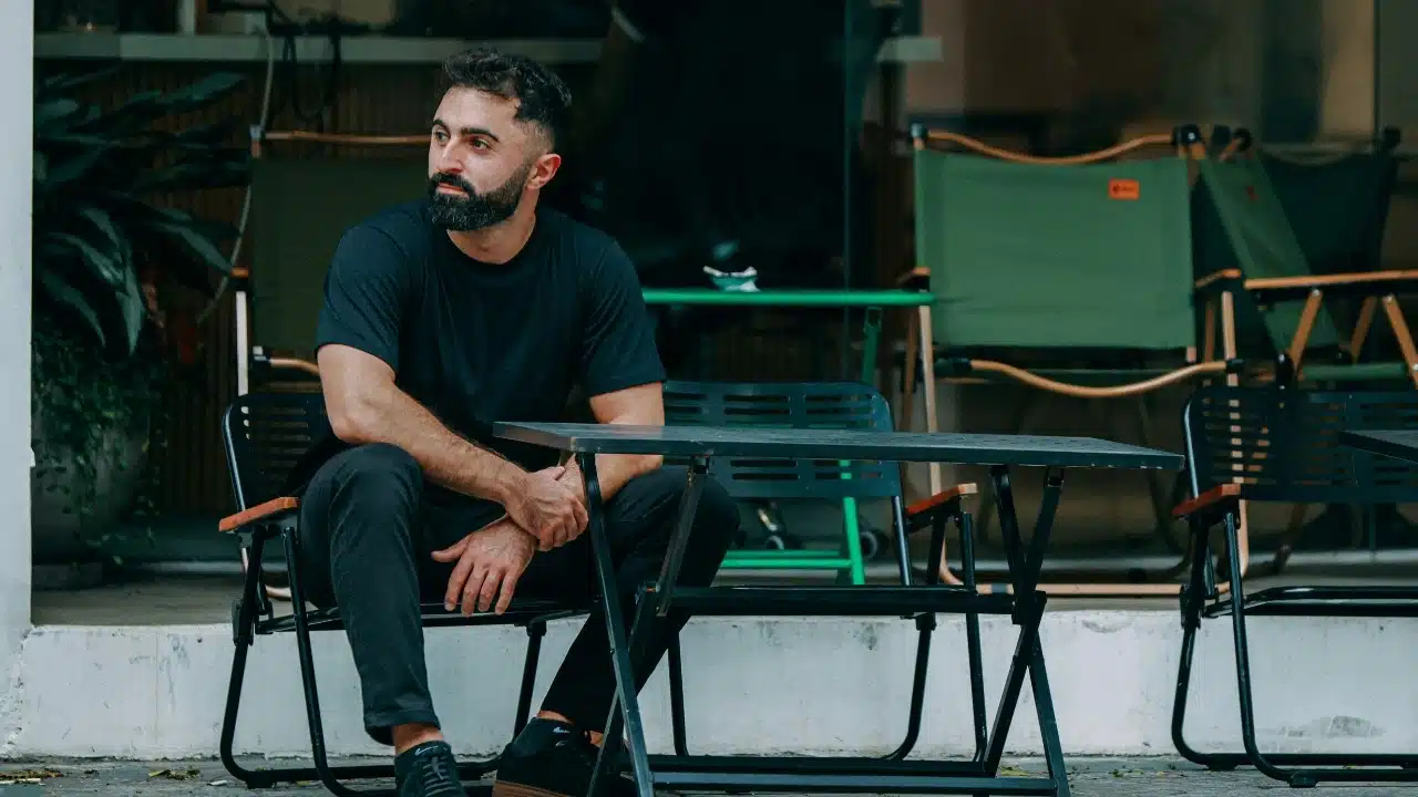 Bearded man in a black t-shirt sits at an outdoor table looking to the side.
