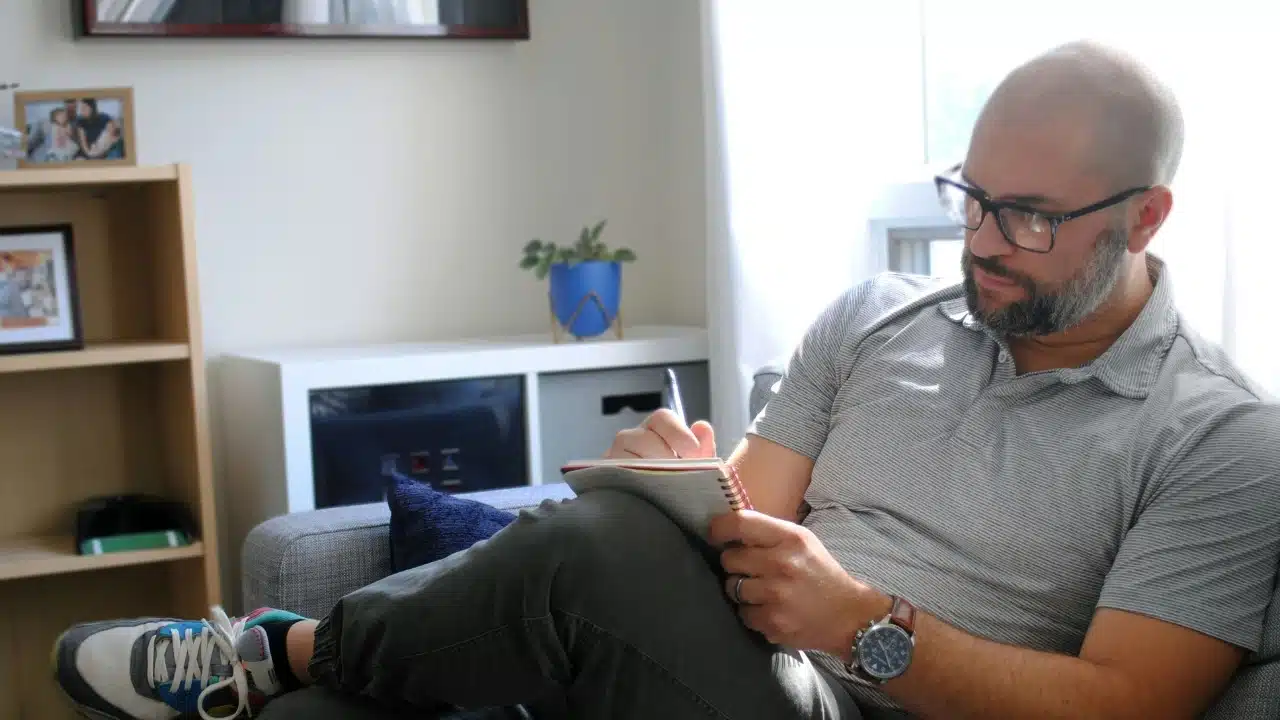 A man with glasses and a beard writes in a small notebook while sitting indoors.
