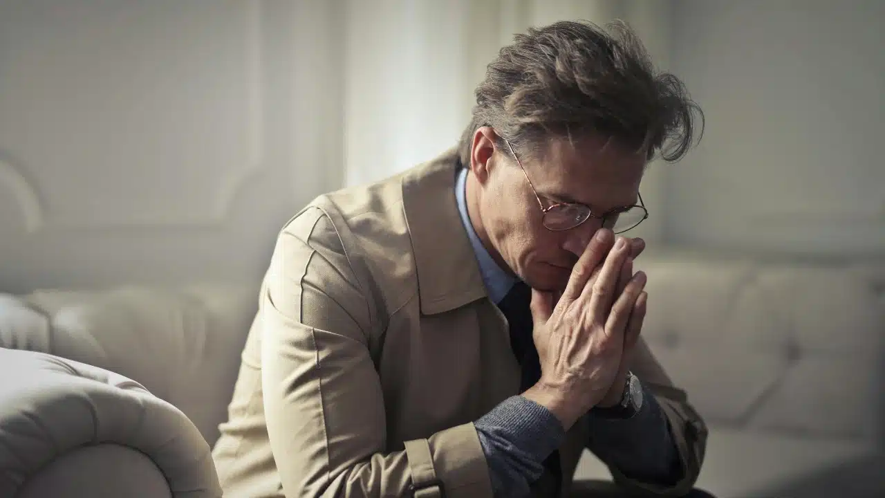 Man in a tan coat sits with hands pressed together near his face, looking down.