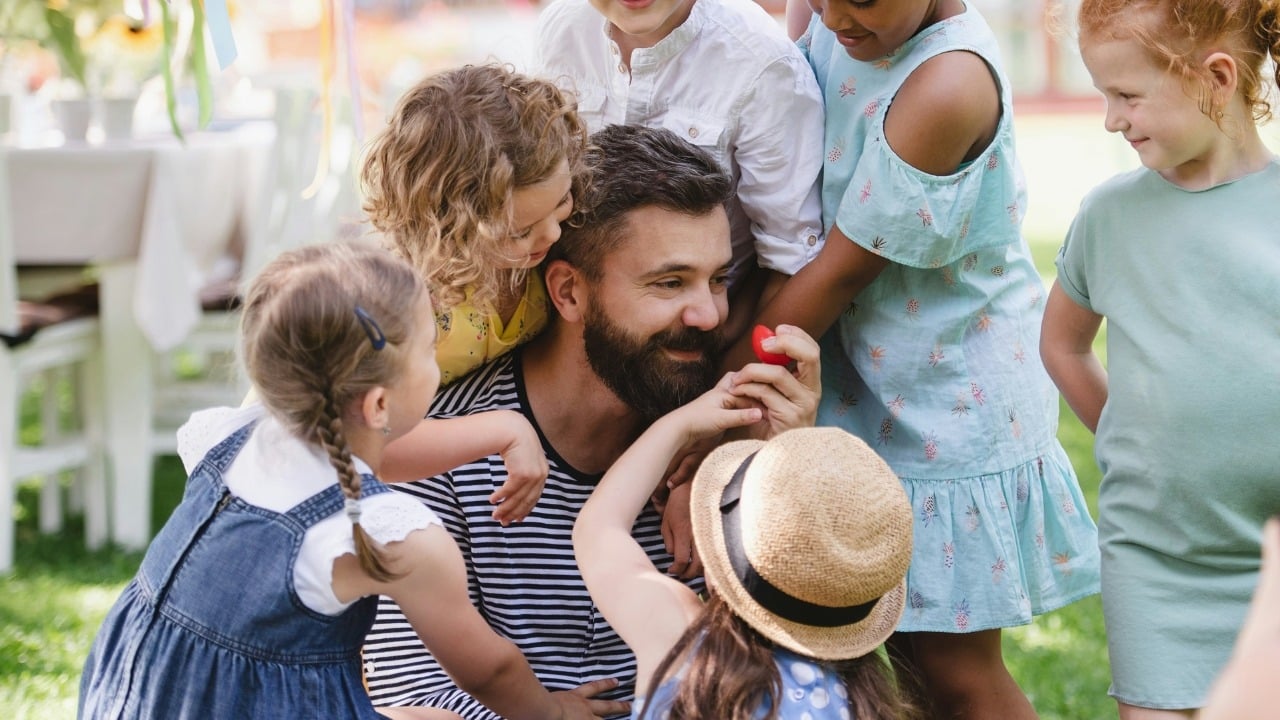 Bearded man in a striped shirt surrounded by several children in an outdoor garden setting.
