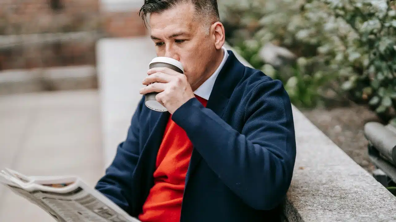 Man in a blue coat and red shirt drinking coffee while reading a newspaper outdoors.