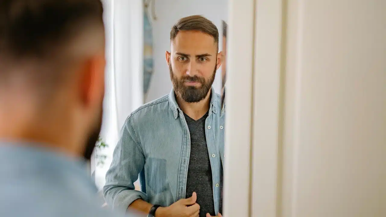 A bearded man in a denim shirt looking at his own reflection in a mirror.