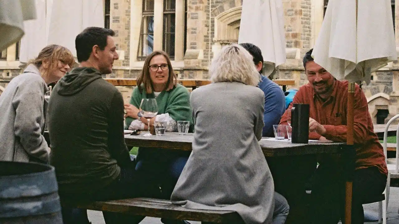 Group of people sitting at a long wooden table outdoors, talking and drinking wine together.