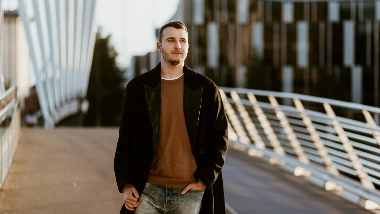 Young man in a long dark coat walking across a modern bridge during the day.