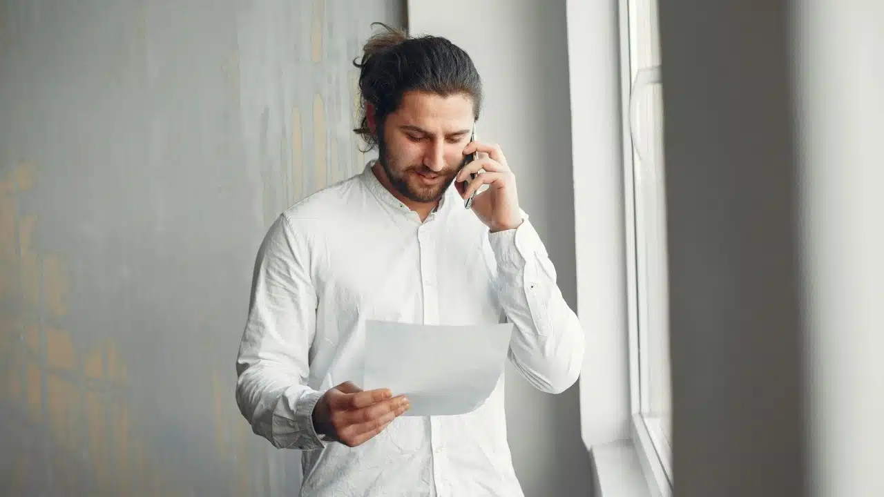 Man with long hair tied back talking on a cell phone while holding a paper.
