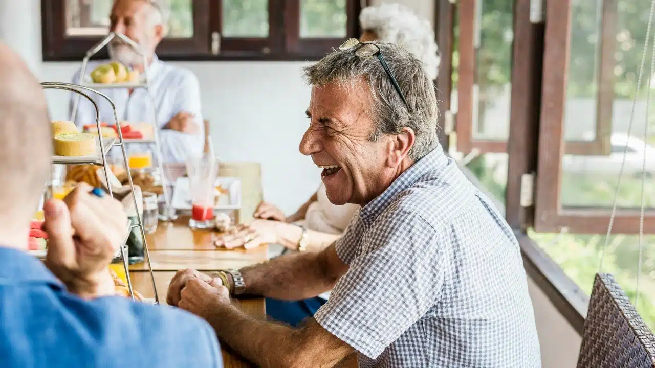 Older man in a checkered shirt laughing loudly at a table with other people.