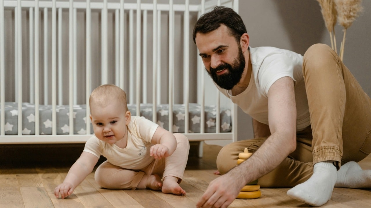 Bearded man sitting on the floor playing with a baby near a white wooden crib.