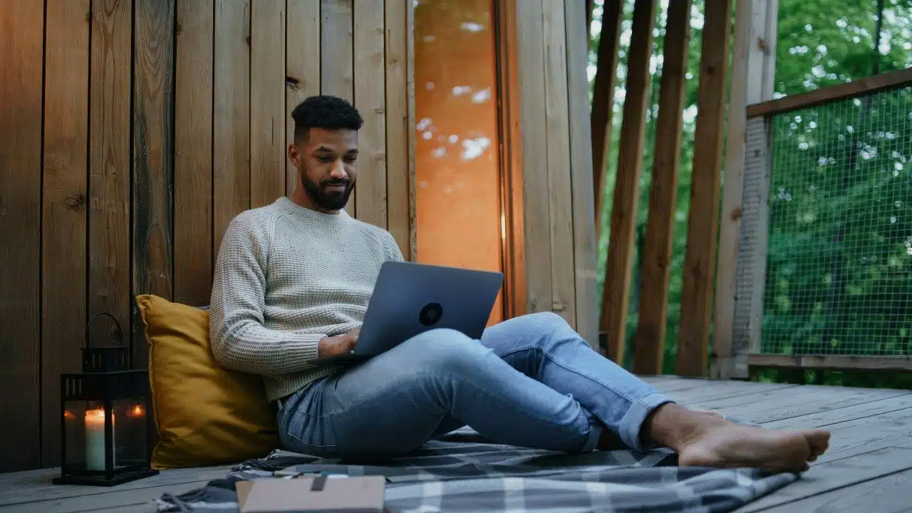 Man sitting on a wooden deck outdoors using a laptop while leaning against a pillow.