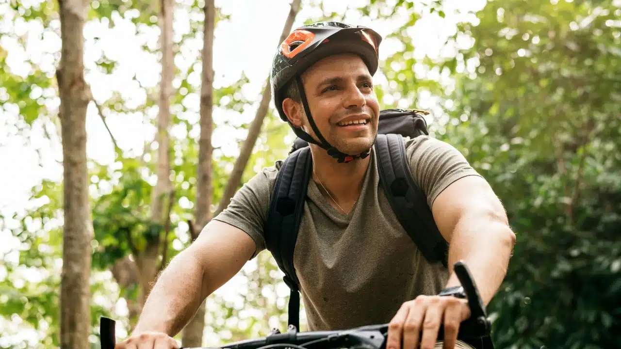 Man wearing a bicycle helmet and backpack smiling while riding through a wooded area outdoors.